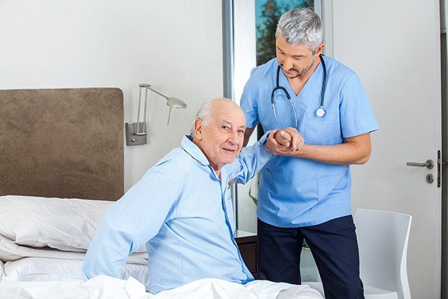 A nurse in blue scrubs helps an elderly man out of bed.