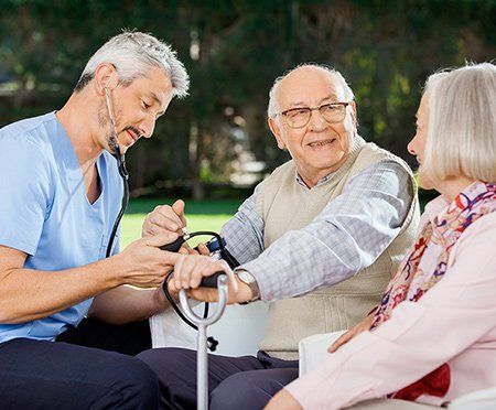 An elderly couple sits in the park while a home health aide takes the man's blood pressure.